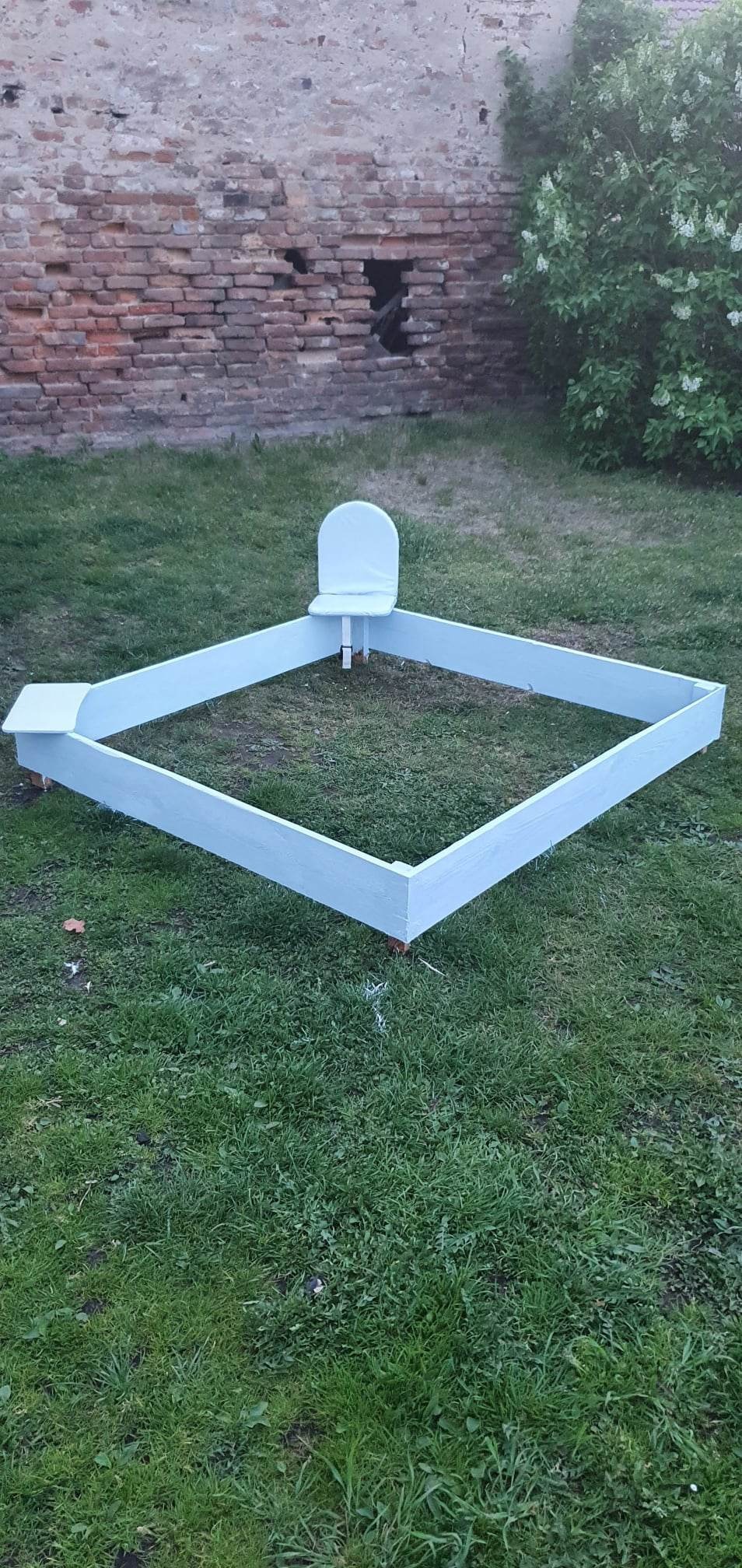 Nisko ustawiona, kwadratowa konstrukcja z jasnych desek z małym krzesełkiem wewnątrz, prawdopodobnie przygotowana jako piaskownica, na trawniku w Łączniku, z ceglaną ścianą i kwitnącym krzewem w tle.