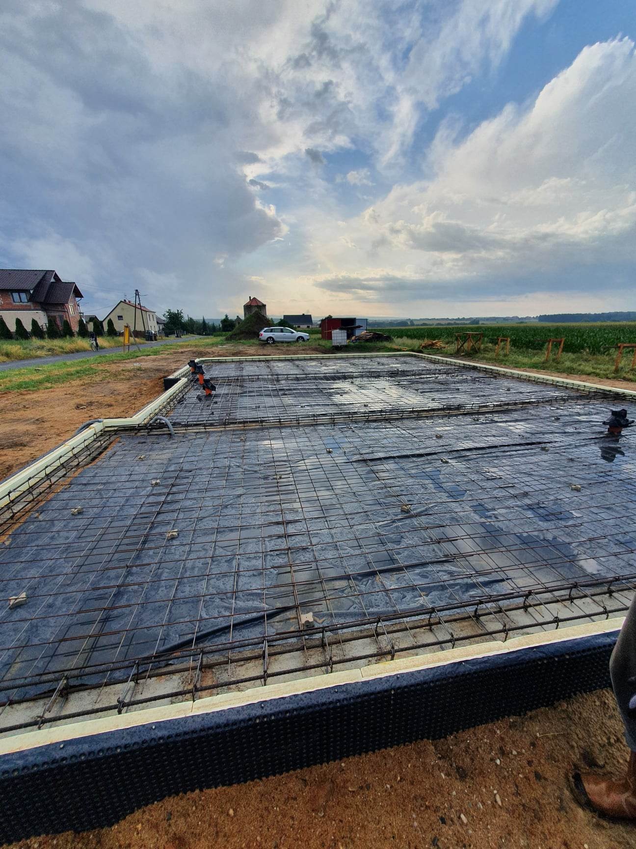 Przygotowanie zbrojenia fundamentu domu na czarnej folii budowlanej, widoczne pręty zbrojeniowe, izolacja termiczna pionowa i pozioma, w tle domy i pochmurne niebo.