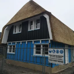 Dom w trakcie ocieplania elewacji, widoczna warstwa niebieskiej izolacji, dach kryty strzechą, ogrodzenie budowy z tablicą informacyjną.