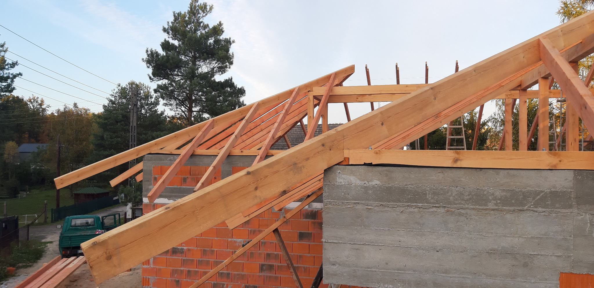 Wznoszenie konstrukcji drewnianej dachu na budynku z czerwonej cegły i szarego betonu, widok na las w tle.