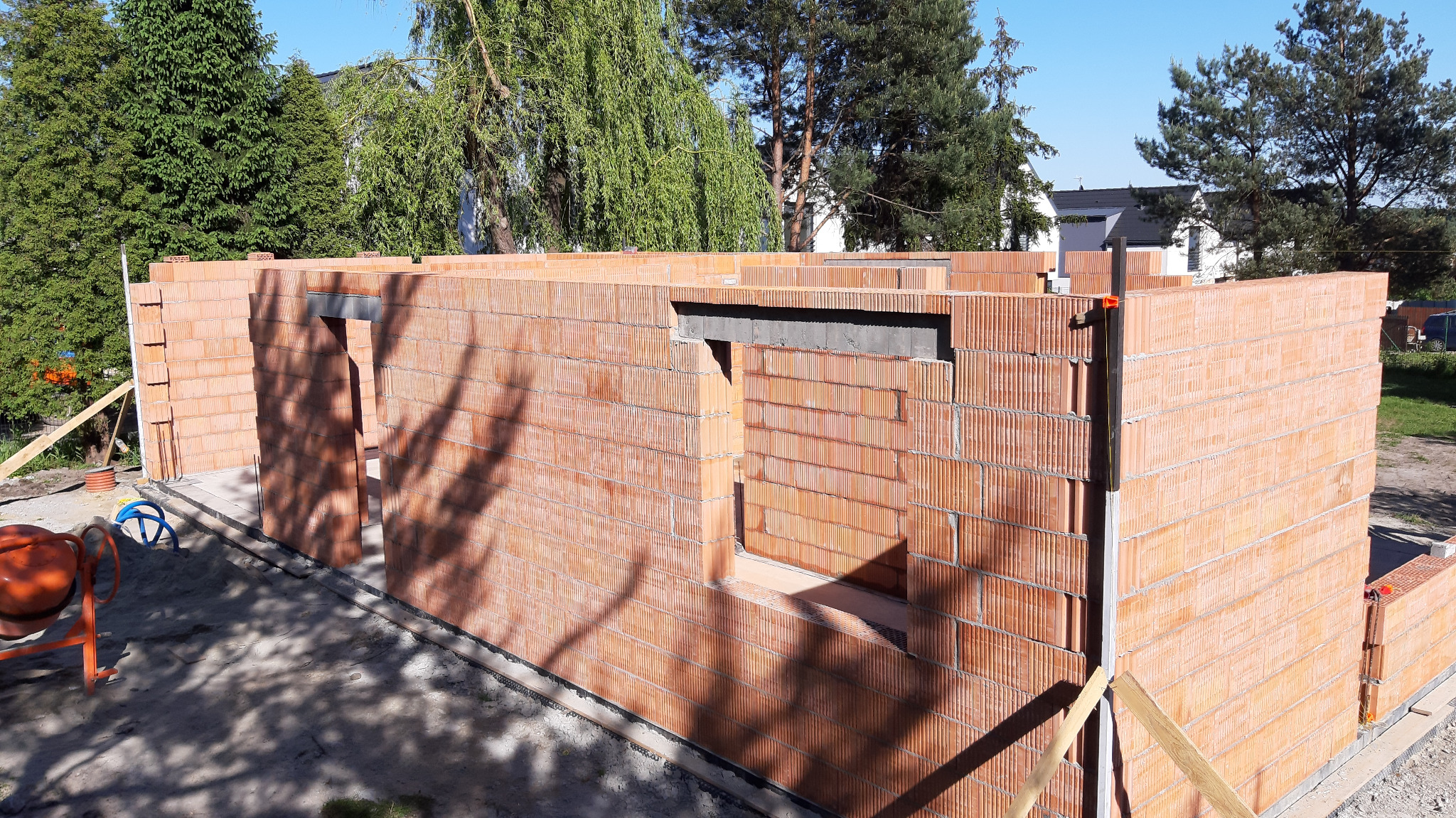 Wznoszenie ścian zewnętrznych z czerwonej ceramiki poryzowanej, widoczne otwory okienne i drzwiowe, na zewnątrz stoi betoniarka.