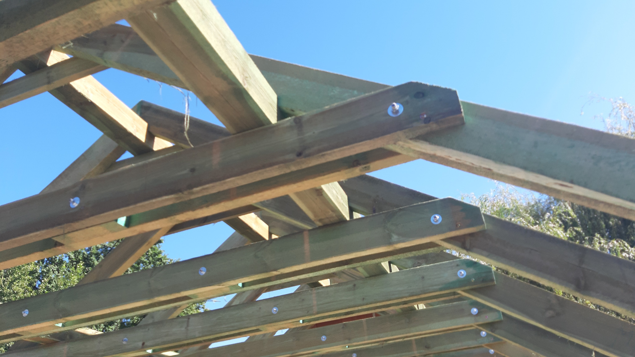 Konstrukcja drewniana dachu z widocznymi łącznikami śrubowymi, świeżo zaimpregnowana na tle błękitnego nieba.