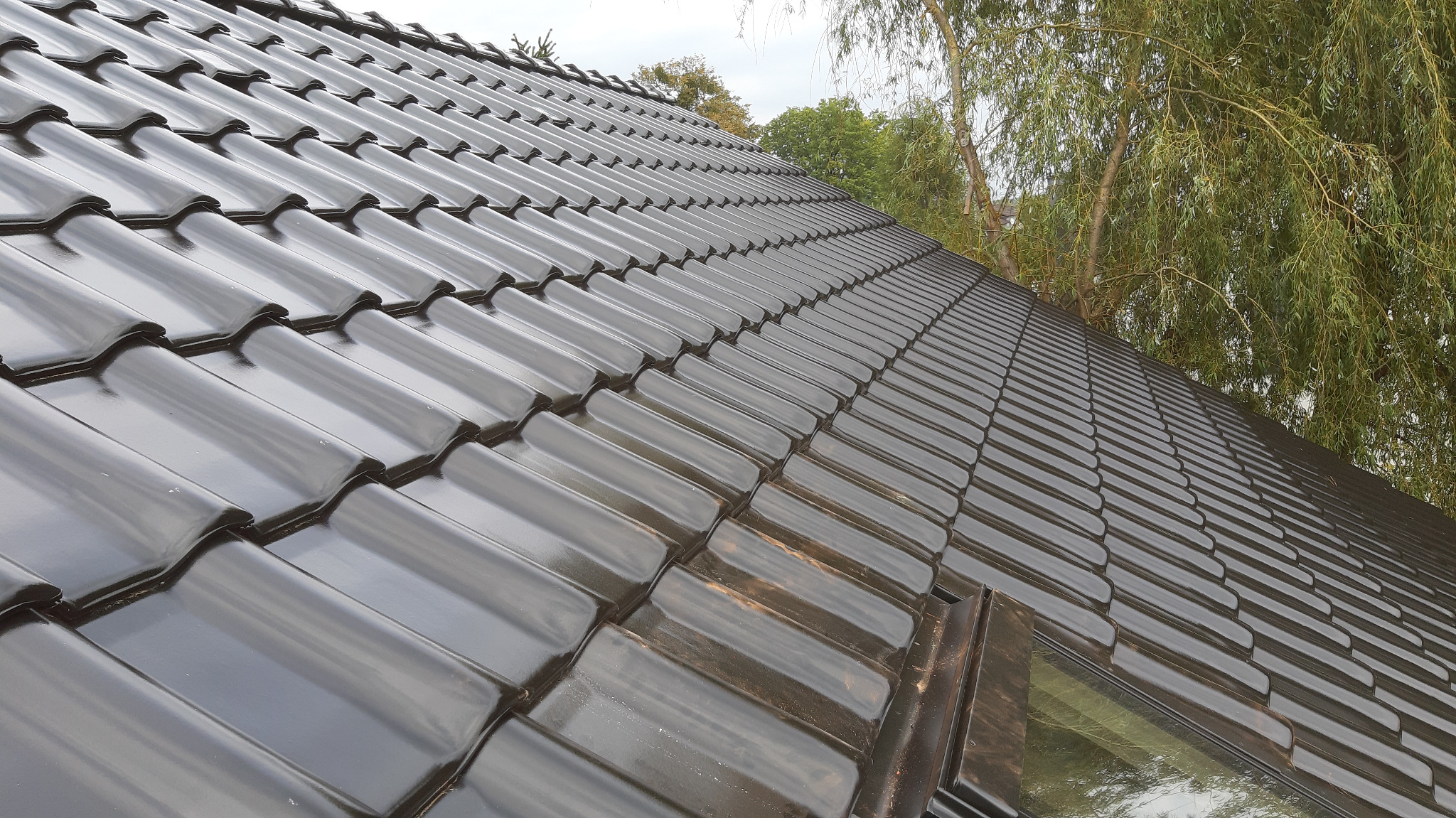 Błyszczące, nowe, ciemnobrązowe dachówki ceramiczne ułożone na dachu z widocznym oknem dachowym i fragmentami drzew w tle.