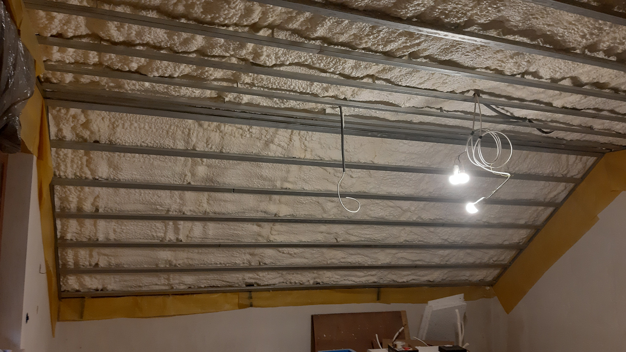 Poddasze ocieplone pianką poliuretanową między metalowymi profilami, widoczne przewody elektryczne i żarówki tymczasowego oświetlenia.
