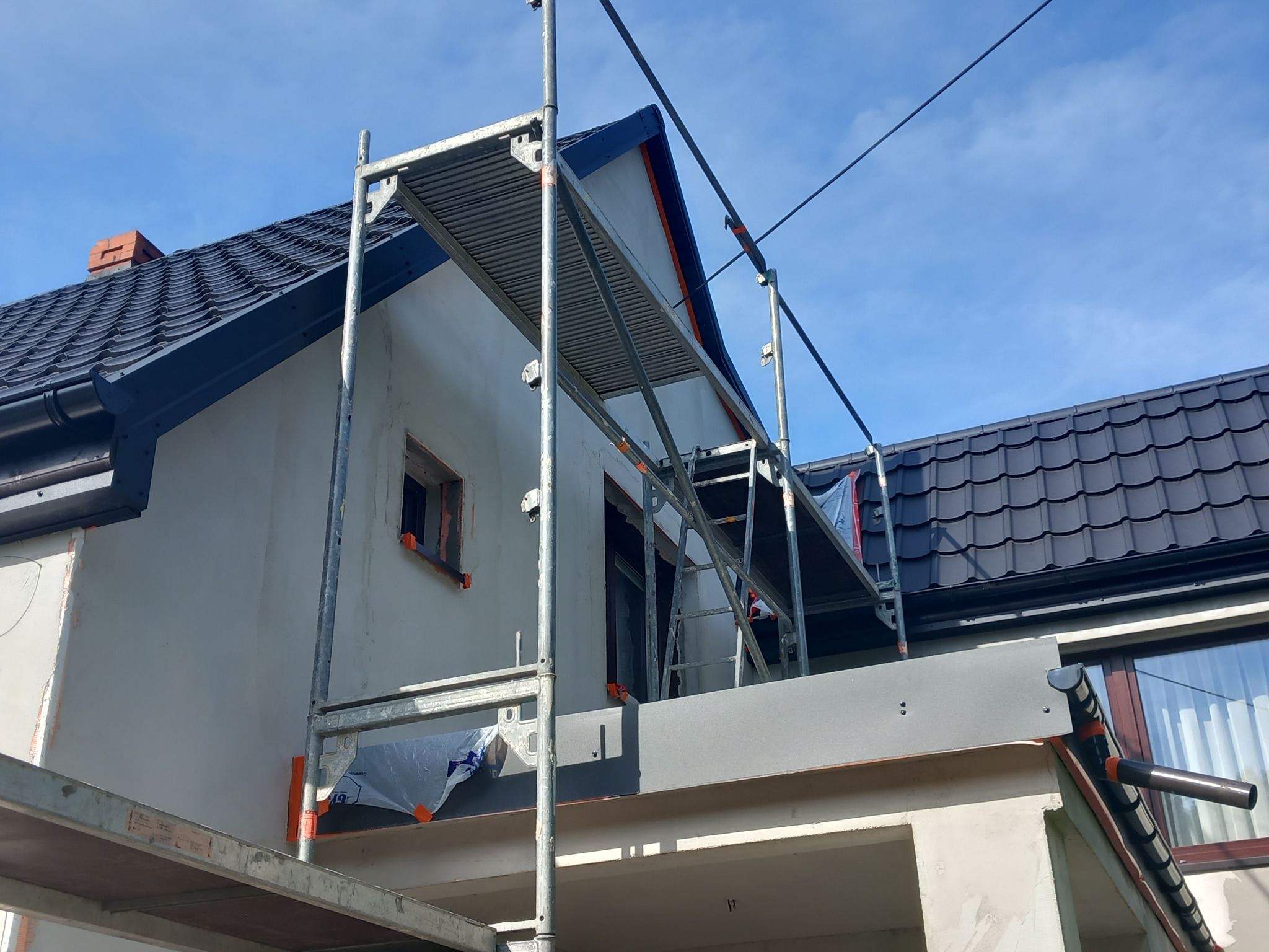 Dom jednorodzinny w trakcie rozbudowy, widoczne rusztowania, fragment dachu z dachówką, ocieplone ściany oraz przygotowane otwory okienne.