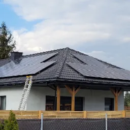 Dom jednorodzinny z panelami słonecznymi na dachu w Tomaszowie Mazowieckim, drabina oparta o ścianę, widoczne ogrodzenie i drzewa w tle.