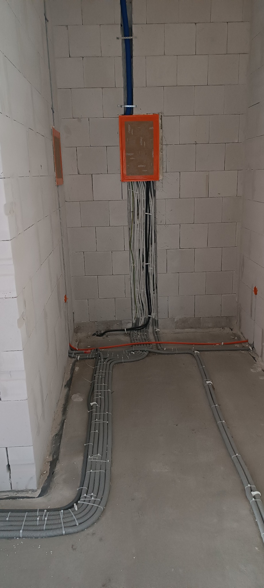 Surowe wnętrze z widocznymi przewodami elektrycznymi w ścianach i podłodze, zabezpieczonymi pomarańczową taśmą osłonową i szarymi rurami karbowanymi, z białymi ścianami z bloczków betonowych.