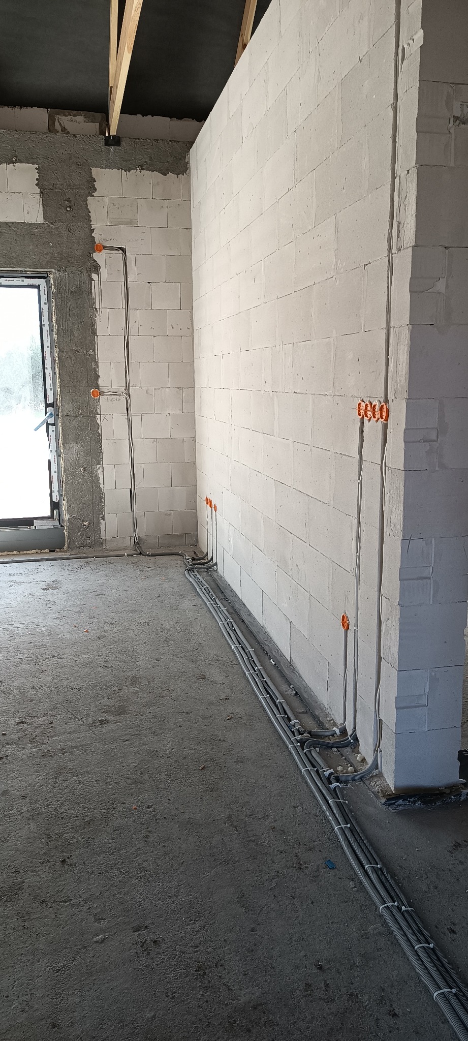 Surowy stan wnętrza z widocznymi przewodami elektrycznymi w szarych peszlach ułożonymi wzdłuż ściany z białych bloczków, pomarańczowe puszki instalacyjne w ścianie, sufit z drewnianymi belkami.