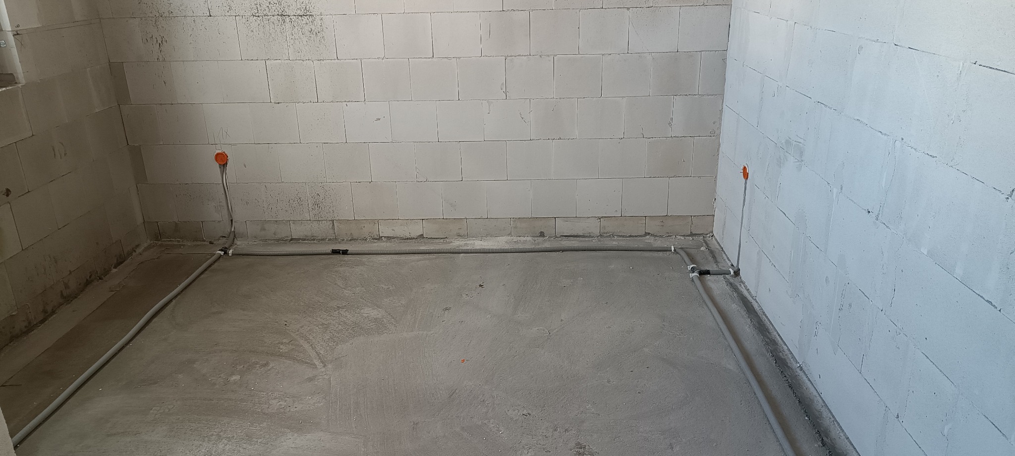 Surowy stan pomieszczenia z widocznymi przewodami elektrycznymi ułożonymi wzdłuż ścian i wyprowadzonymi gniazdkami podtynkowymi, ściany z bloczków betonowych, posadzka betonowa.