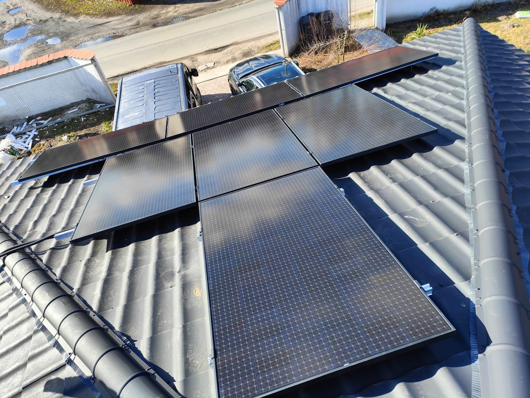 Panele słoneczne zamontowane na szarym dachu z widocznym samochodem osobowym i dostawczym w tle, uchwycone z góry.