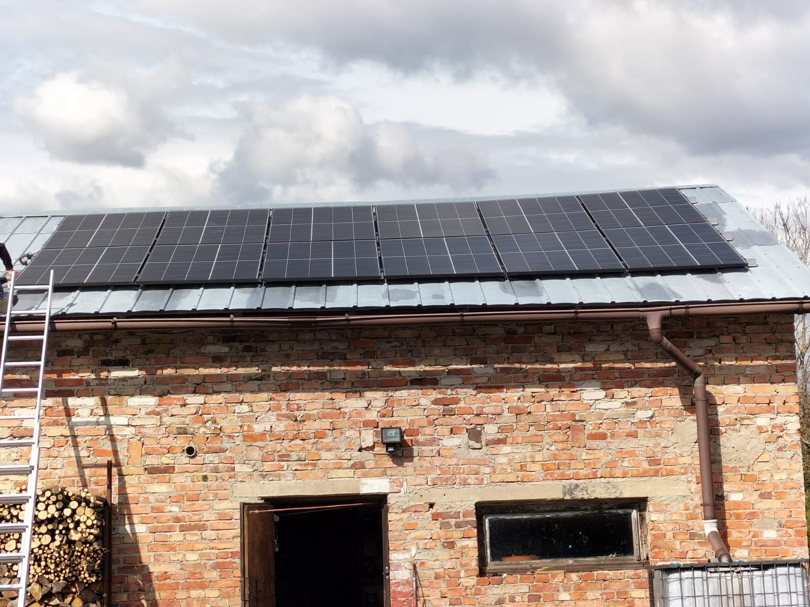 Montaż paneli słonecznych na dachu ceglanego budynku gospodarczego, widoczna drabina i stos drewna przy ścianie.