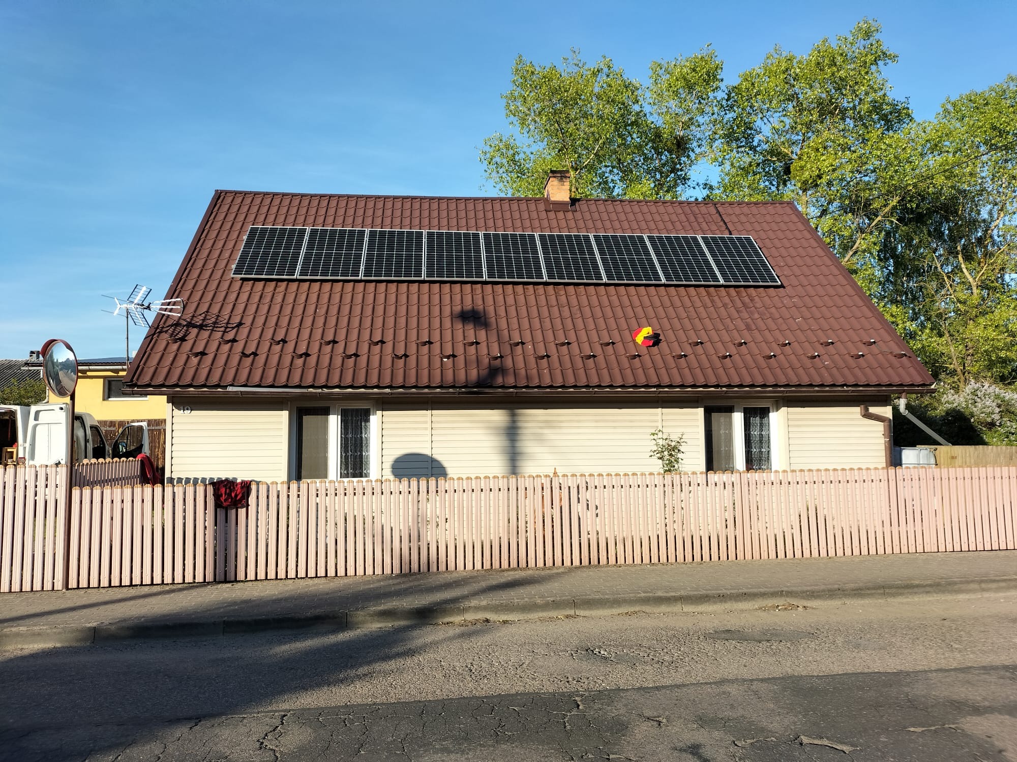 Dom jednorodzinny z brązowym dachem pokrytym panelami słonecznymi, widoczna antena satelitarna, różowe ogrodzenie i drzewa w tle.