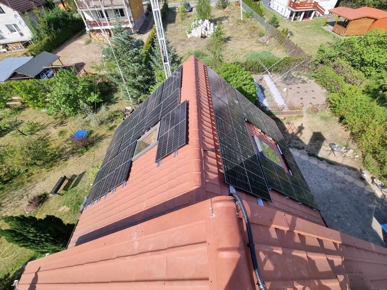 Widok z góry na dach z terakoty z zamontowanymi panelami słonecznymi, pomiędzy którymi znajdują się okna dachowe. Widoczne elementy montażowe paneli oraz czarny kabel. W tle zabudowa jednorodzinna...