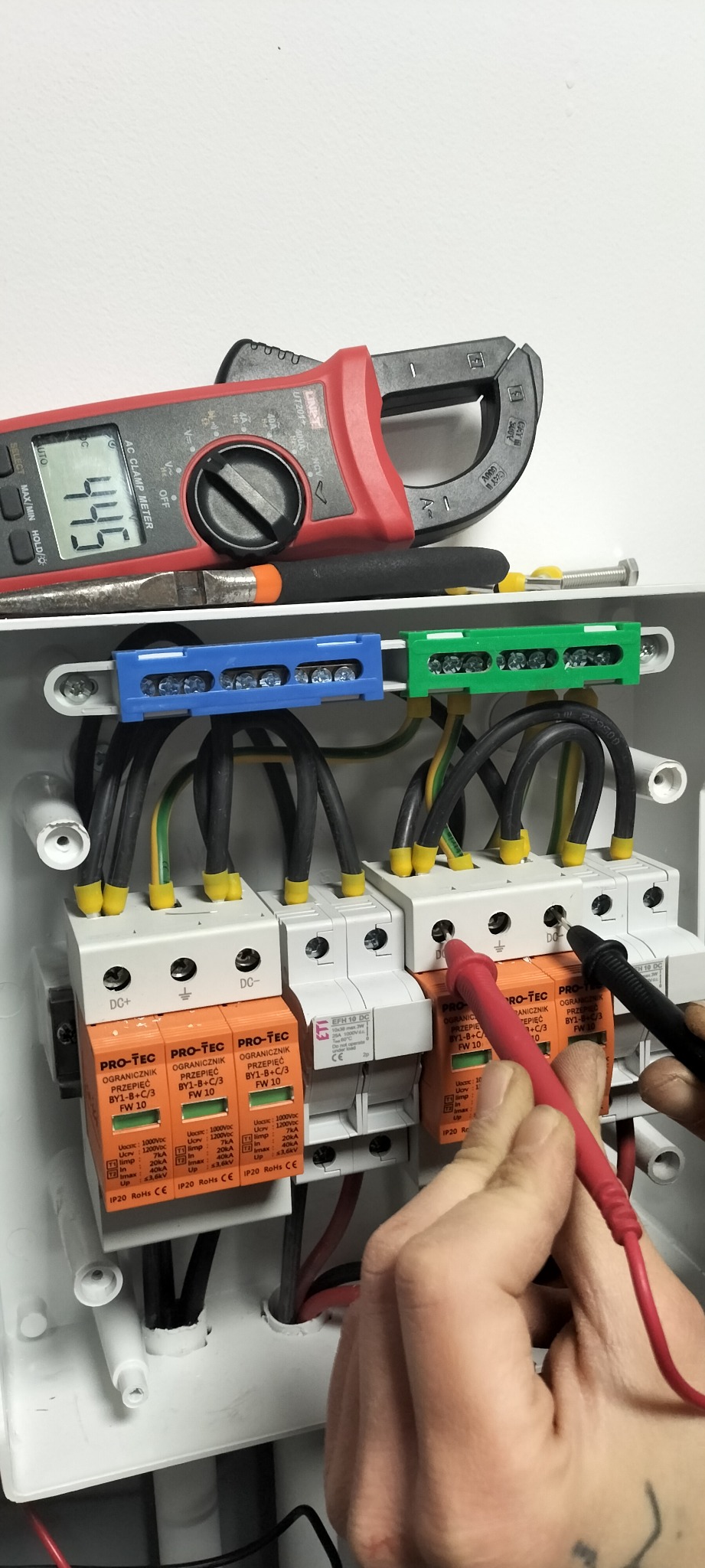 Pomiar napięcia na ogranicznikach przepięć fotowoltaiki w rozdzielnicy, multimetr pokazuje 445V, widoczne listwy zaciskowe i okablowanie.