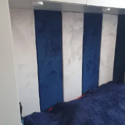 Tapicerowany zagłówek łóżka w pionowe pasy: granatowy welur i szary beton architektoniczny, wkomponowany między białe szafki z oświetleniem punktowym.