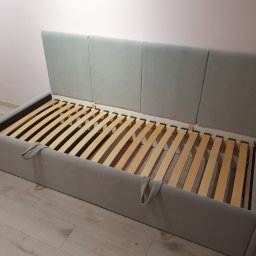 Meble na wymiar - Szary tapicerowany stelaż łóżka z drewnianym rusztem, widoczne uchwyty z tkaniny, tło stanowią ściana i podłoga w odcieniach beżu.