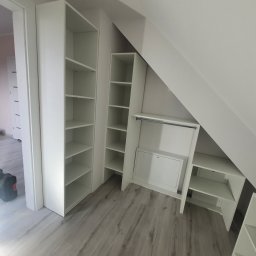 Meble na wymiar - Białe, modułowe regały i szafki wnękowe na poddaszu, z widocznymi narzędziami w tle, podkreślające wykorzystanie przestrzeni i funkcjonalność.