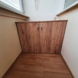 Drewniana szafka z dwoma drzwiczkami, dopasowana do wnęki pod oknem, z widocznym gniazdkiem elektrycznym na ścianie i suszarką na pranie pod sufitem.