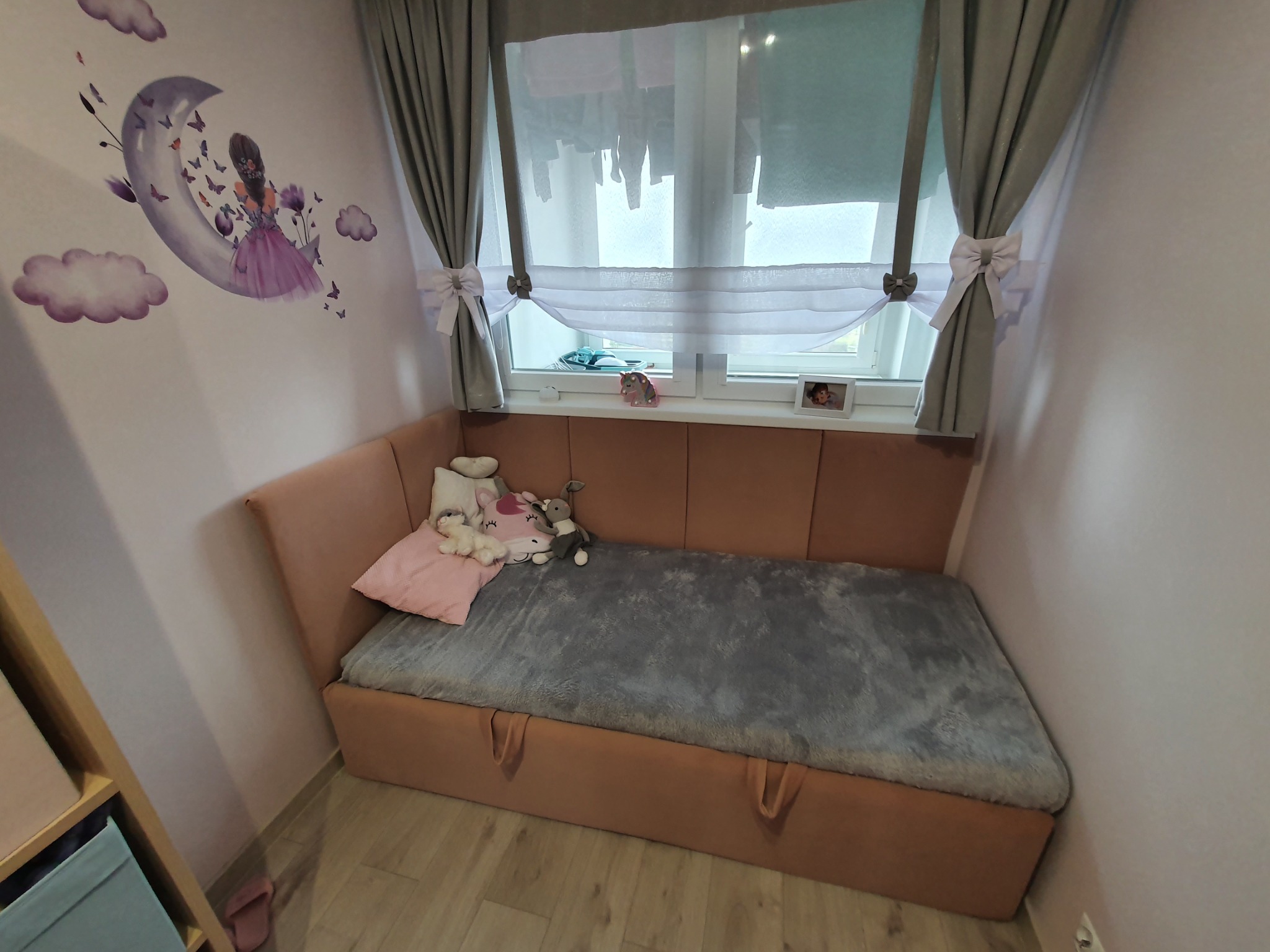 Pokój dziecięcy z łóżkiem w zabudowie, różowe ściany z motywem księżyca i dziewczynki, szare zasłony z kokardami przy oknie.