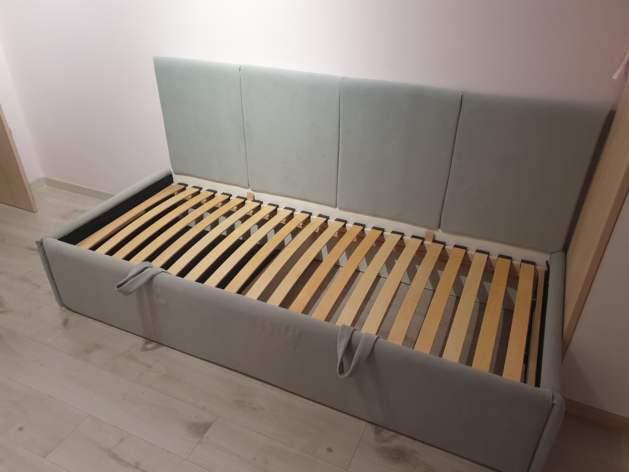 Szare łóżko tapicerowane z drewnianym stelażem listwowym, widoczne uchwyty po bokach, tło stanowią ściany w odcieniach bieli i beżu.