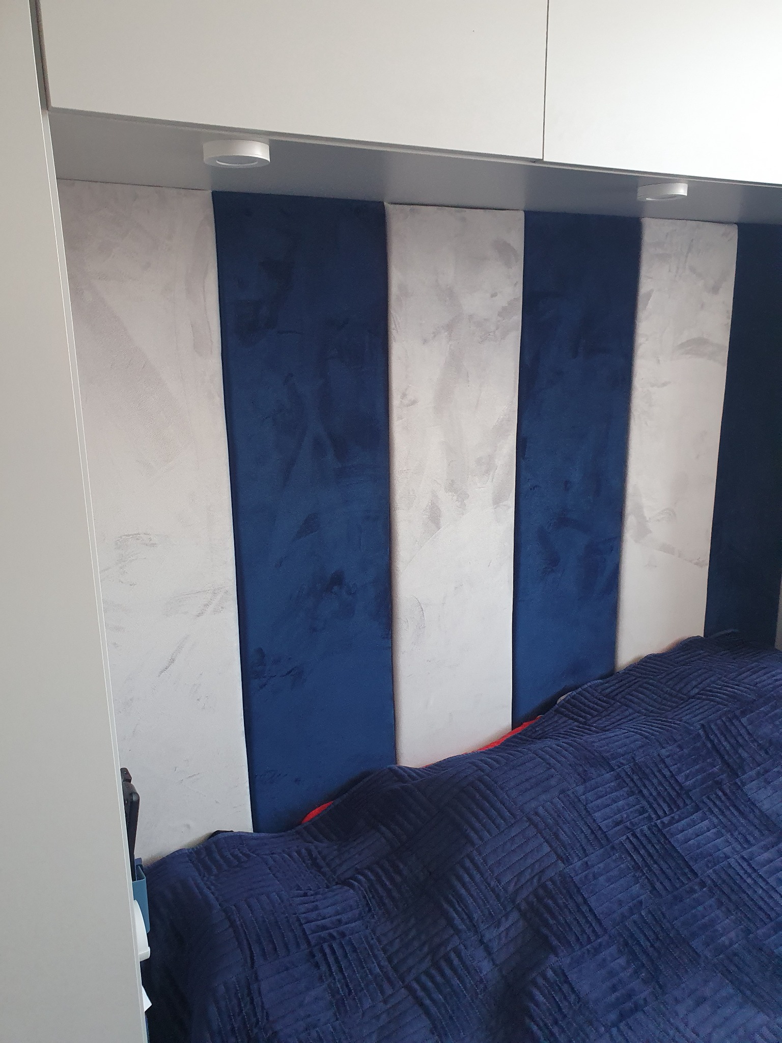 Tapicerowany zagłówek łóżka w pionowe pasy: granatowy welur i szary beton architektoniczny, wkomponowany między białe szafki z oświetleniem punktowym.