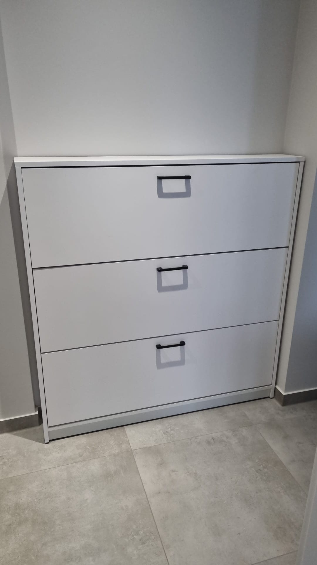 Trzypoziomowa, biała szafka na buty z czarnymi, prostokątnymi uchwytami, umieszczona w przedpokoju na szarej podłodze z płytek.