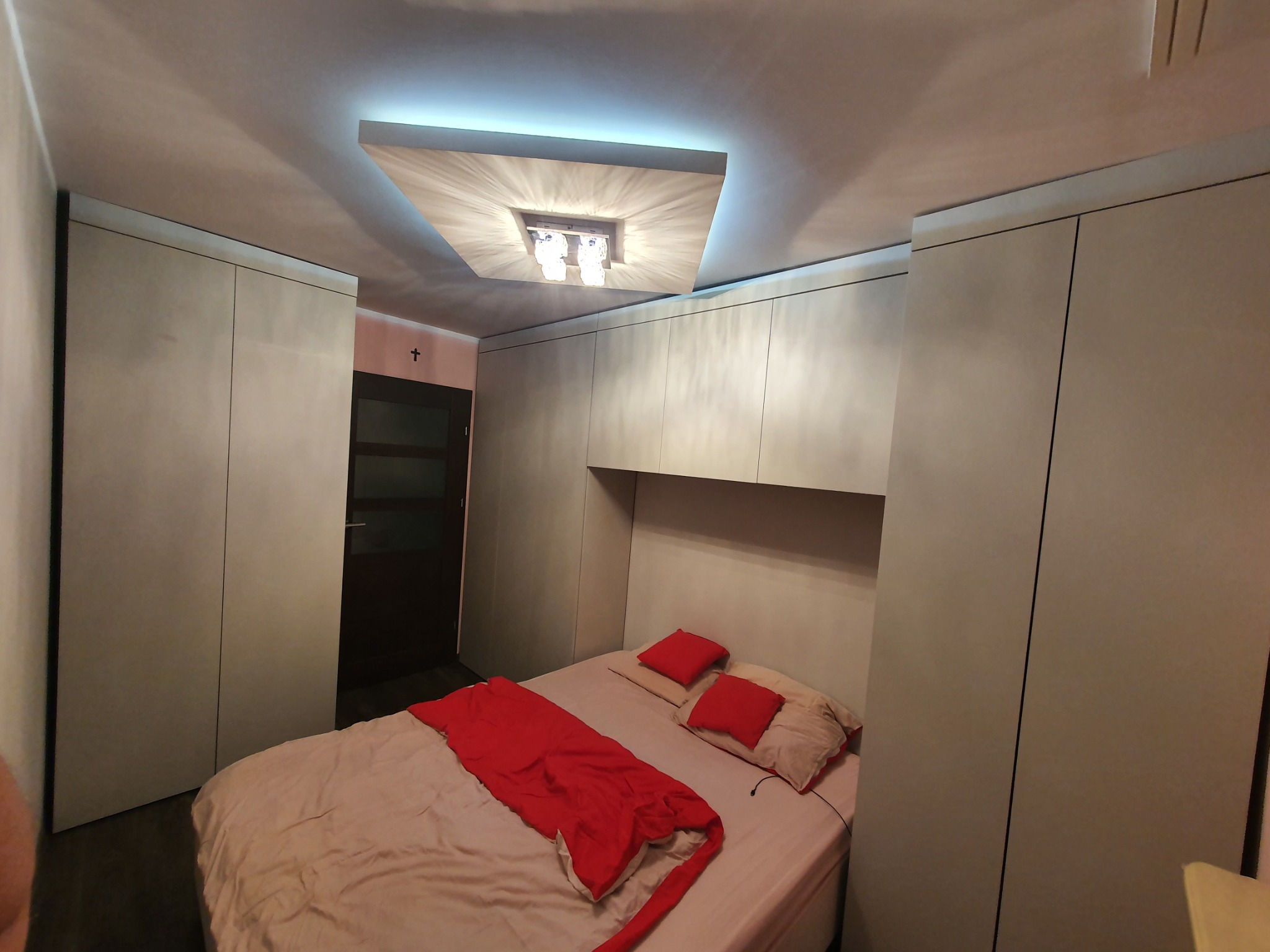 Jasna sypialnia z łóżkiem, czerwonym pledem i szafami wnękowymi po bokach, oświetlona dekoracyjnym plafonem.