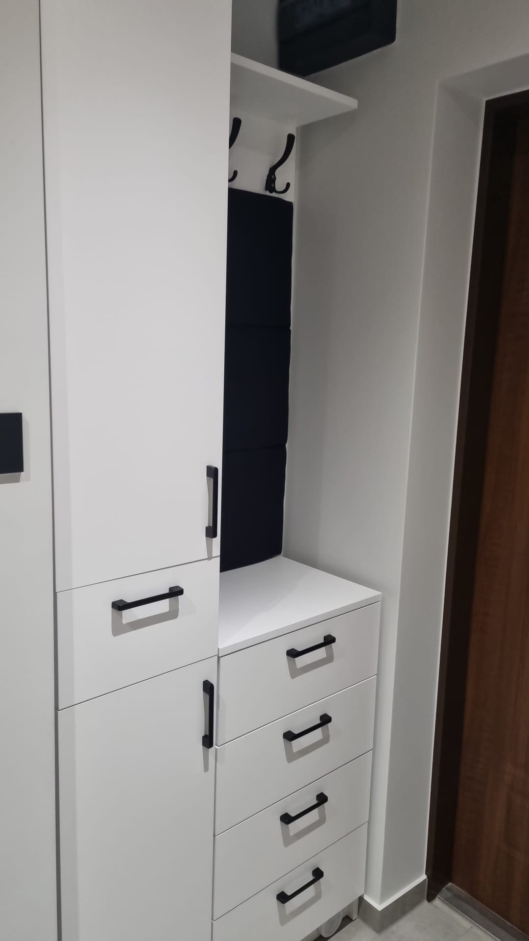 Biała szafa z czarnymi uchwytami, wnęka z haczykami i tapicerowanym panelem, widok z boku przy wejściu.