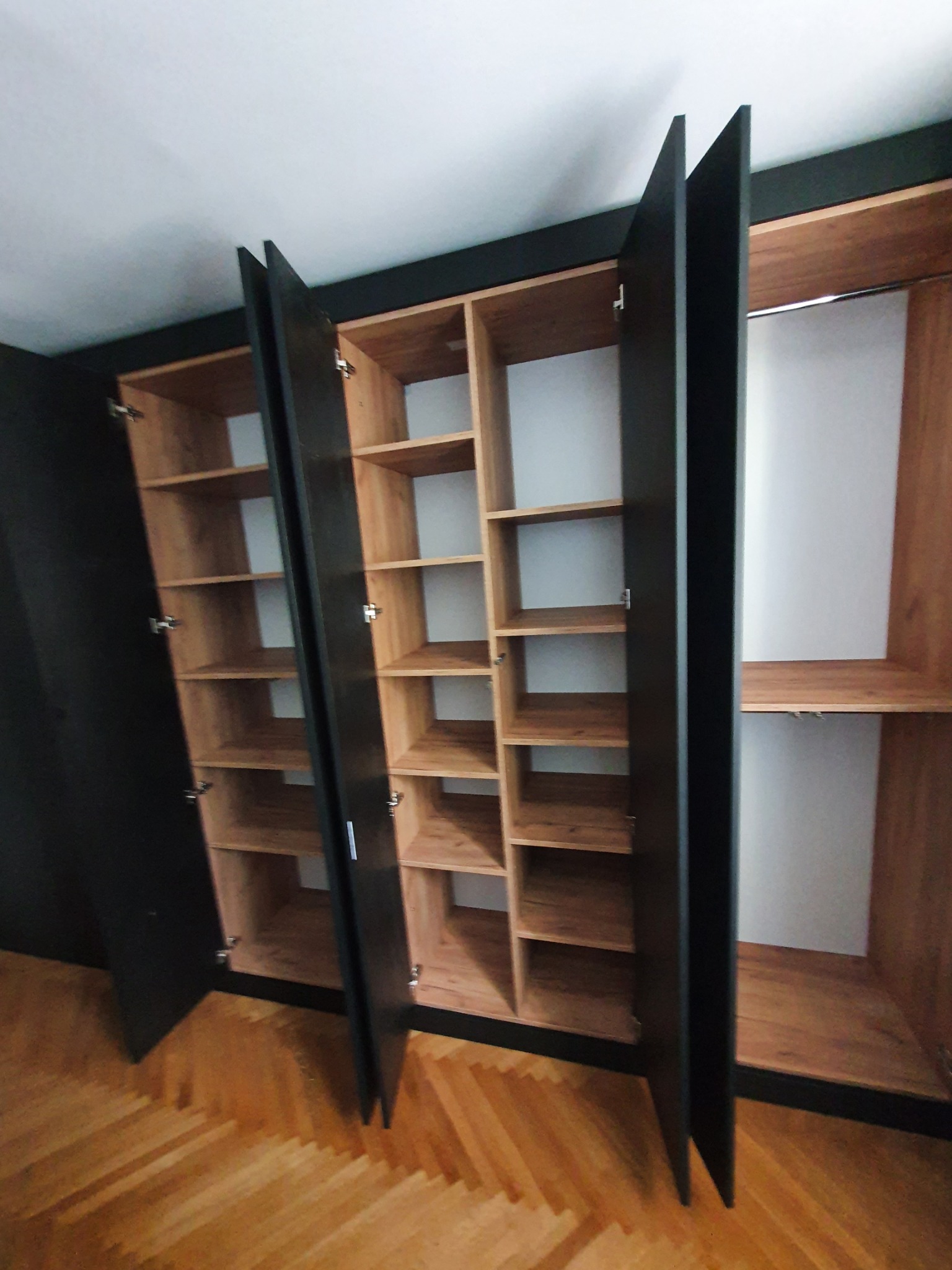 Otwarta, trzydrzwiowa szafa z czarnymi frontami i jasnymi, drewnianymi półkami, widoczna na tle parkietu ułożonego w jodełkę.