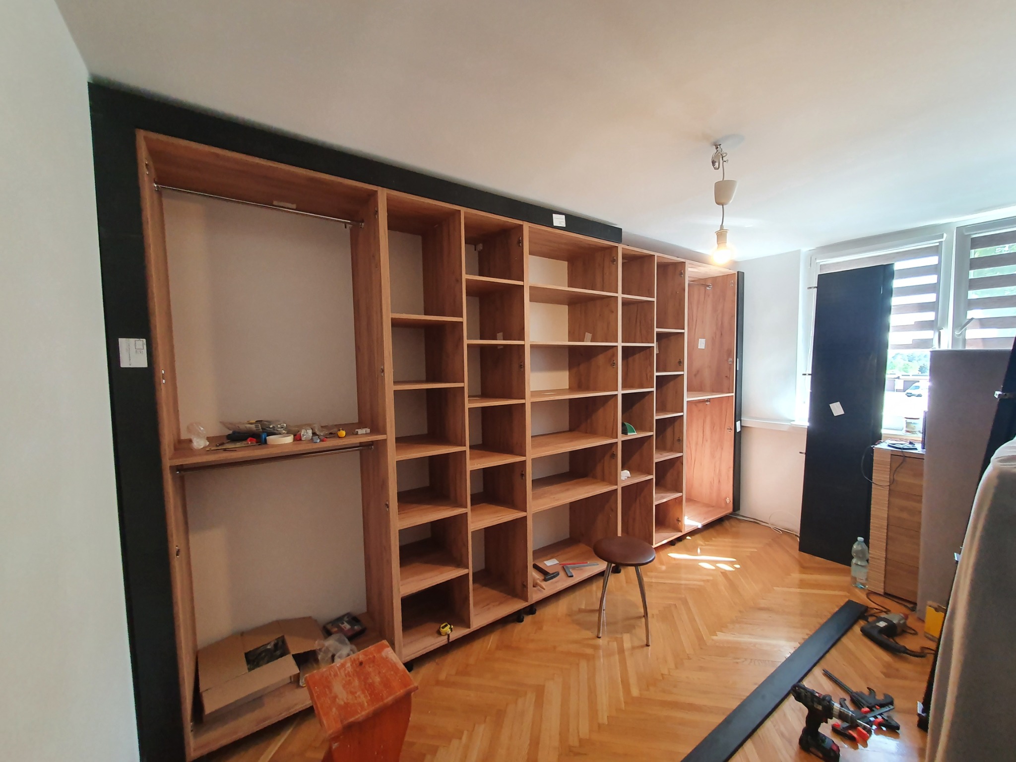 Montaż drewnianej konstrukcji szafy z półkami i drążkami na ubrania, widoczne narzędzia i stołek w jasnym wnętrzu z parkietem.