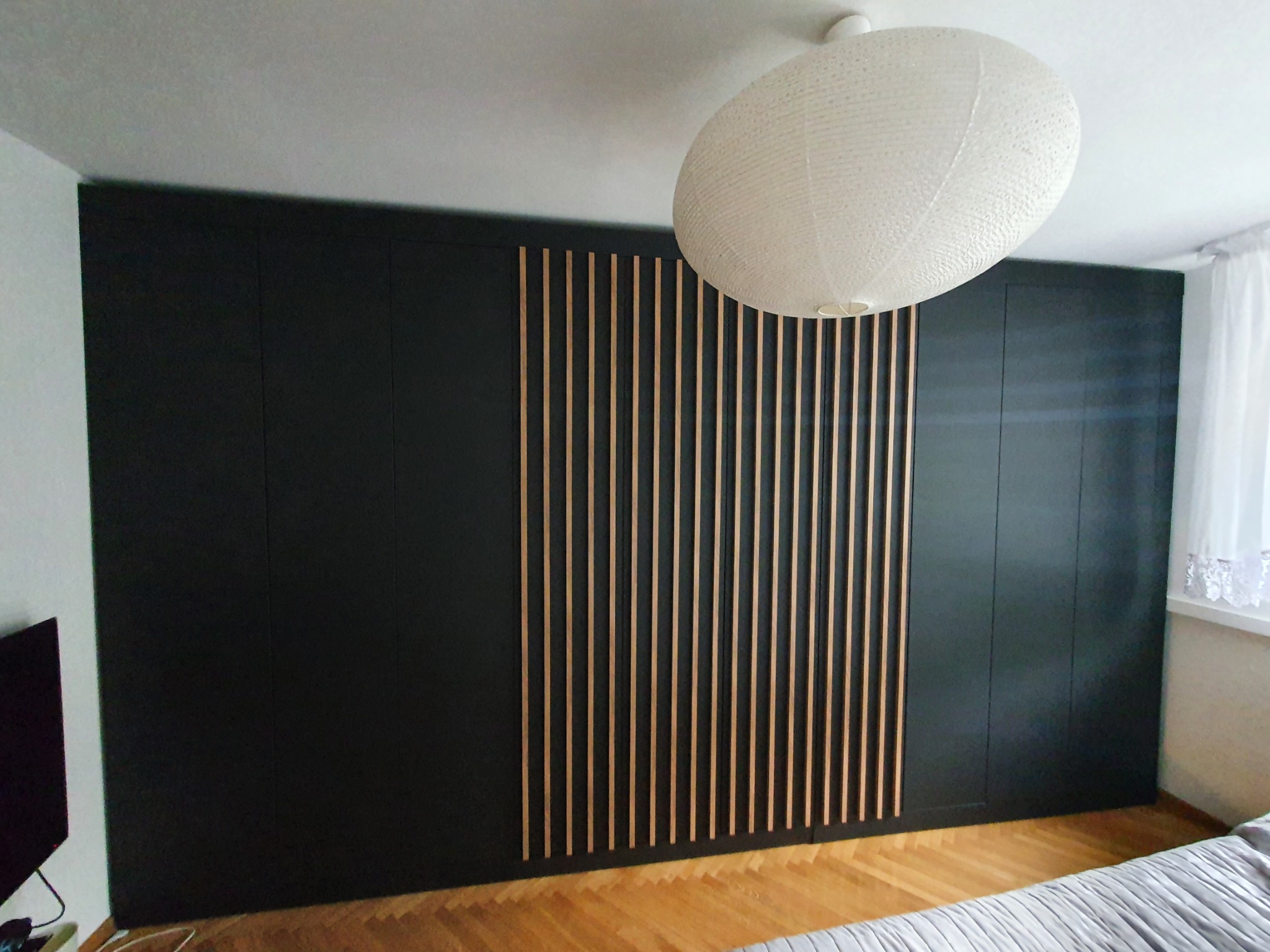 Ciemna zabudowa meblowa z drewnianymi listwami dekoracyjnymi na ścianie, widoczna lampa wisząca i fragment parkietu.