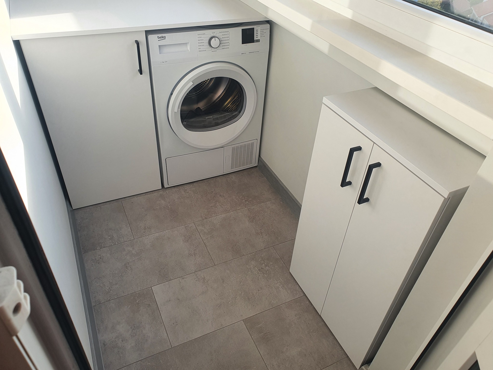 Biała zabudowa z szafkami i pralko-suszarką Beko, minimalistyczne czarne uchwyty, szara podłoga z płytek imitujących kamień, widok z góry.