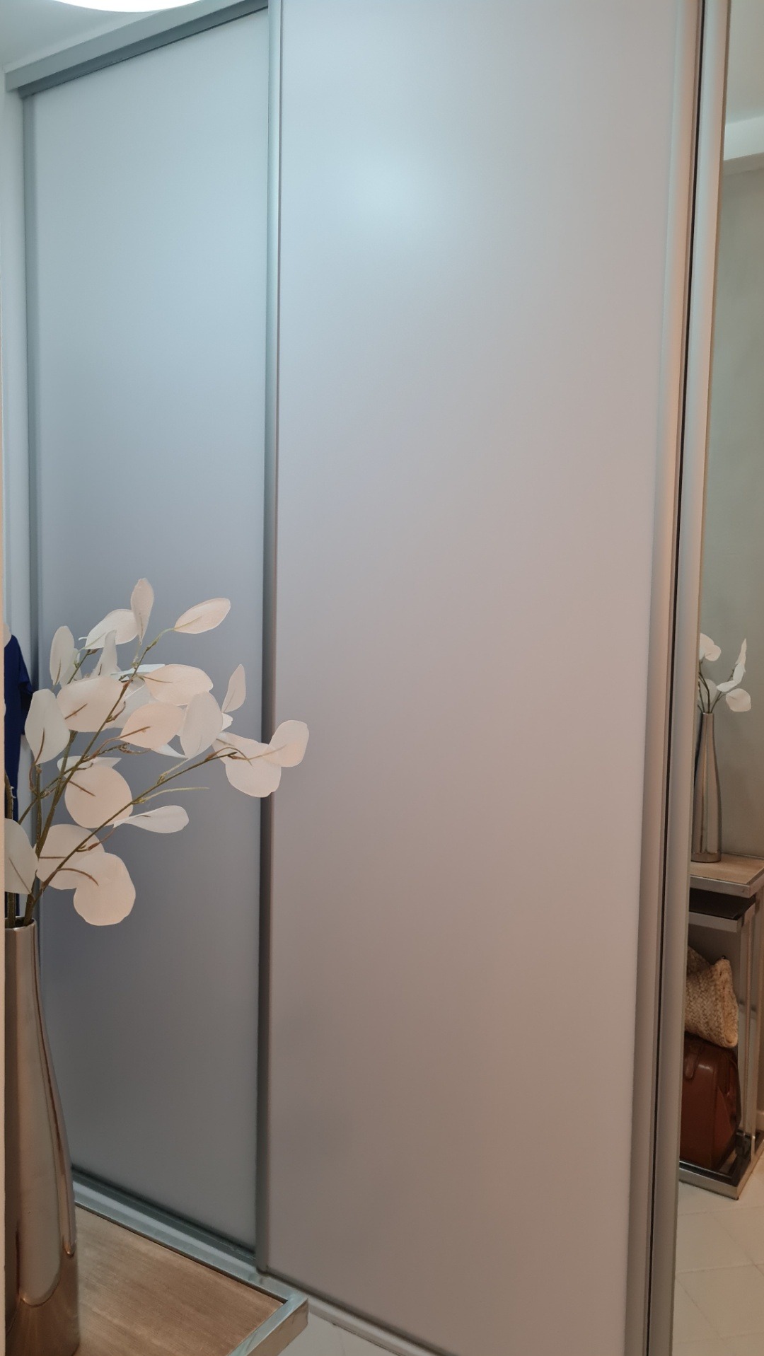 Nowoczesna szafa wnękowa z przesuwnymi drzwiami w kolorze jasnego popielu, z dekoracyjnym wazonem z białymi kwiatami na pierwszym planie, widok z bliska.