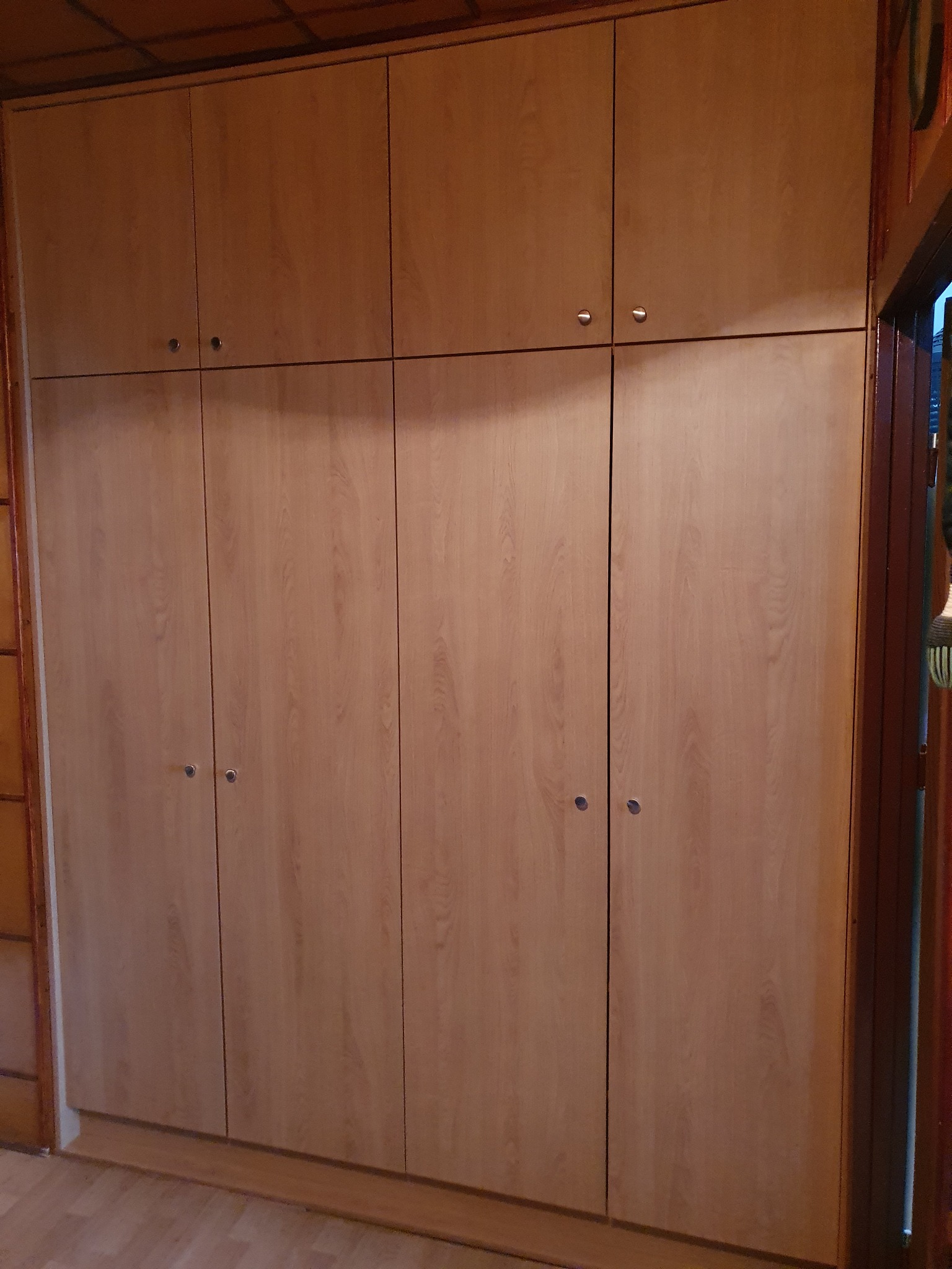 Jasna, drewnopodobna szafa wnękowa z górnymi szafkami, widoczna w korytarzu domu, z małymi, okrągłymi uchwytami.