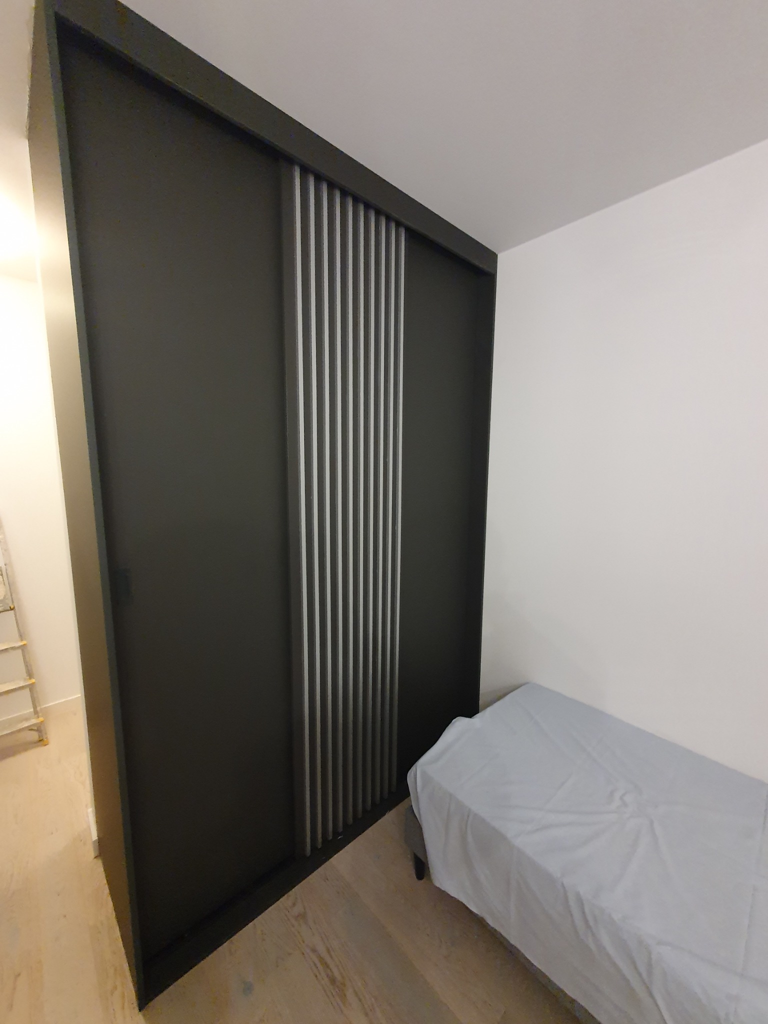 Nowoczesna szafa przesuwna w kolorze grafitowym z dekoracyjnymi pionowymi listwami, umieszczona w sypialni obok łóżka z szarą pościelą.
