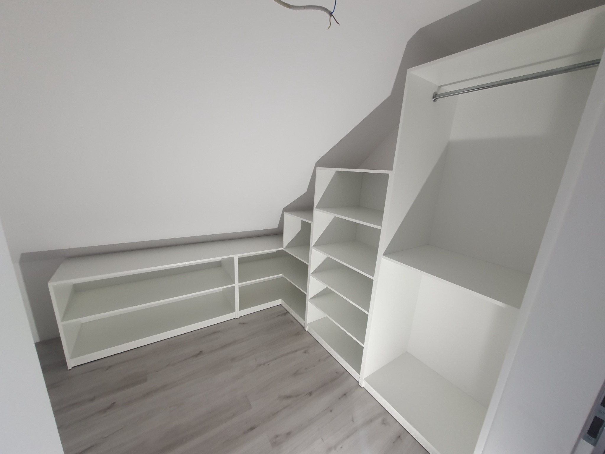 Biała zabudowa wnęki na poddaszu ze skośnym sufitem, półki i drążek na ubrania, szare panele na podłodze, widoczne kable wystające z sufitu.
