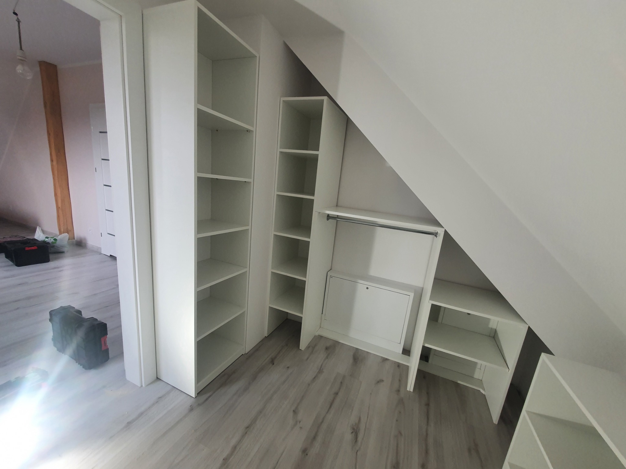 Białe, modułowe regały i szafki wnękowe na poddaszu, z widocznymi narzędziami w tle, podkreślające wykorzystanie przestrzeni i funkcjonalność.