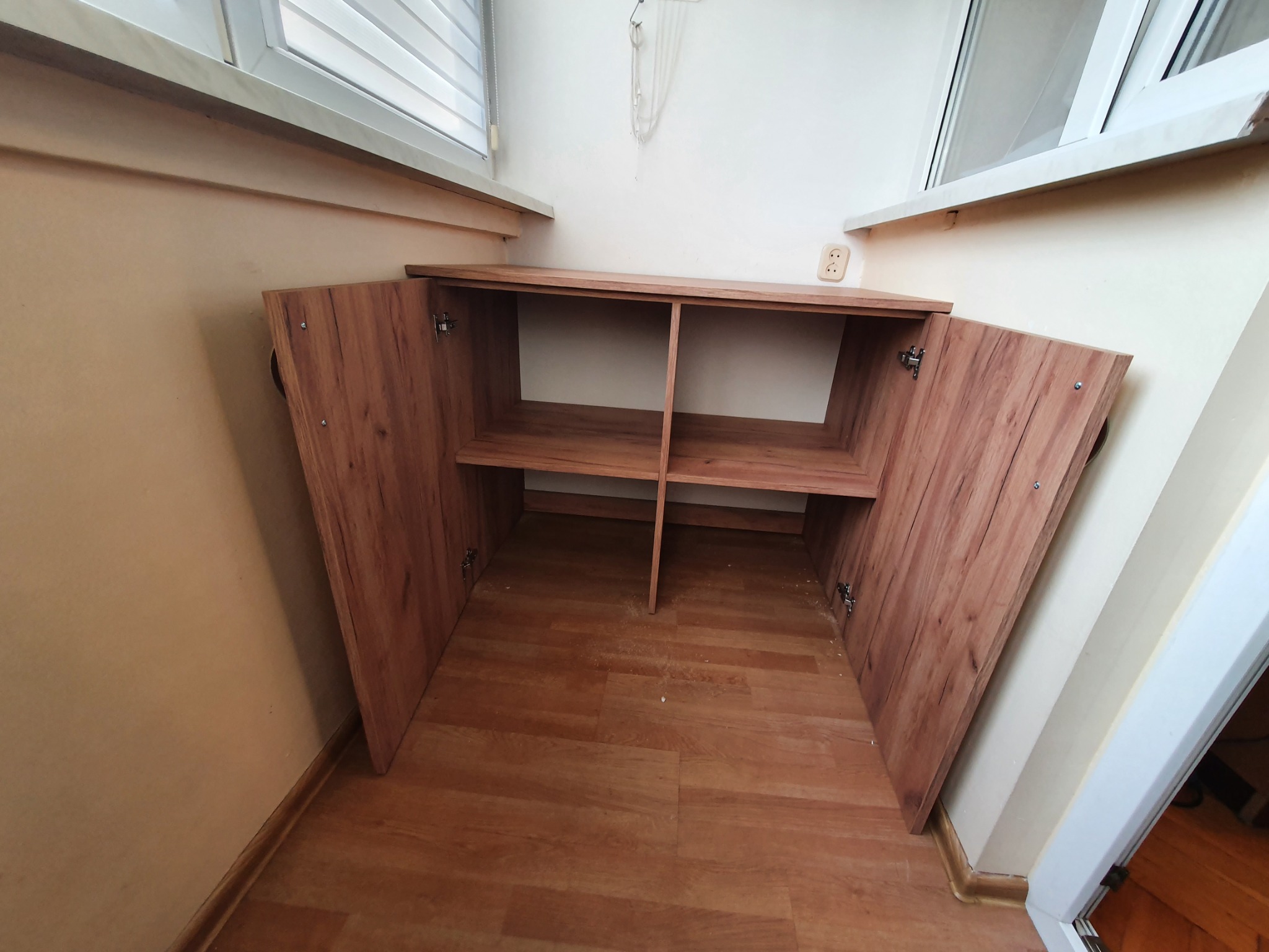 Drewniana szafka z otwartymi drzwiczkami, umieszczona we wnęce między oknem a ścianą, z dwiema półkami wewnątrz, na podłodze z paneli.