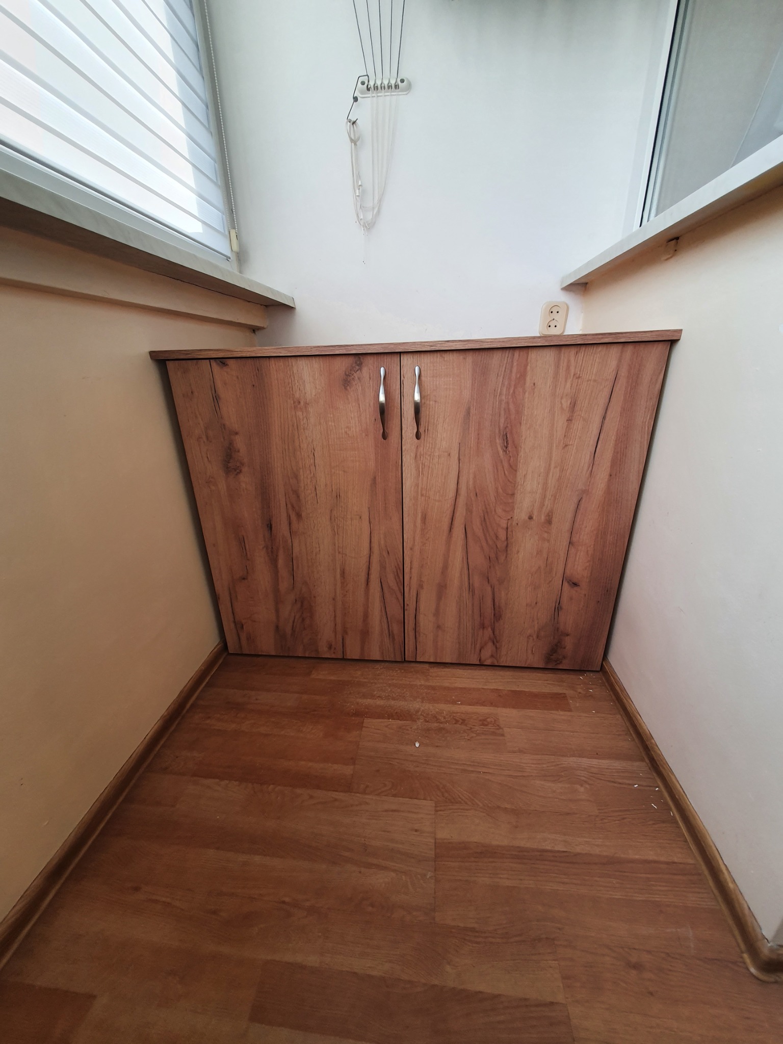 Drewniana szafka z dwoma drzwiczkami, dopasowana do wnęki pod oknem, z widocznym gniazdkiem elektrycznym na ścianie i suszarką na pranie pod sufitem.