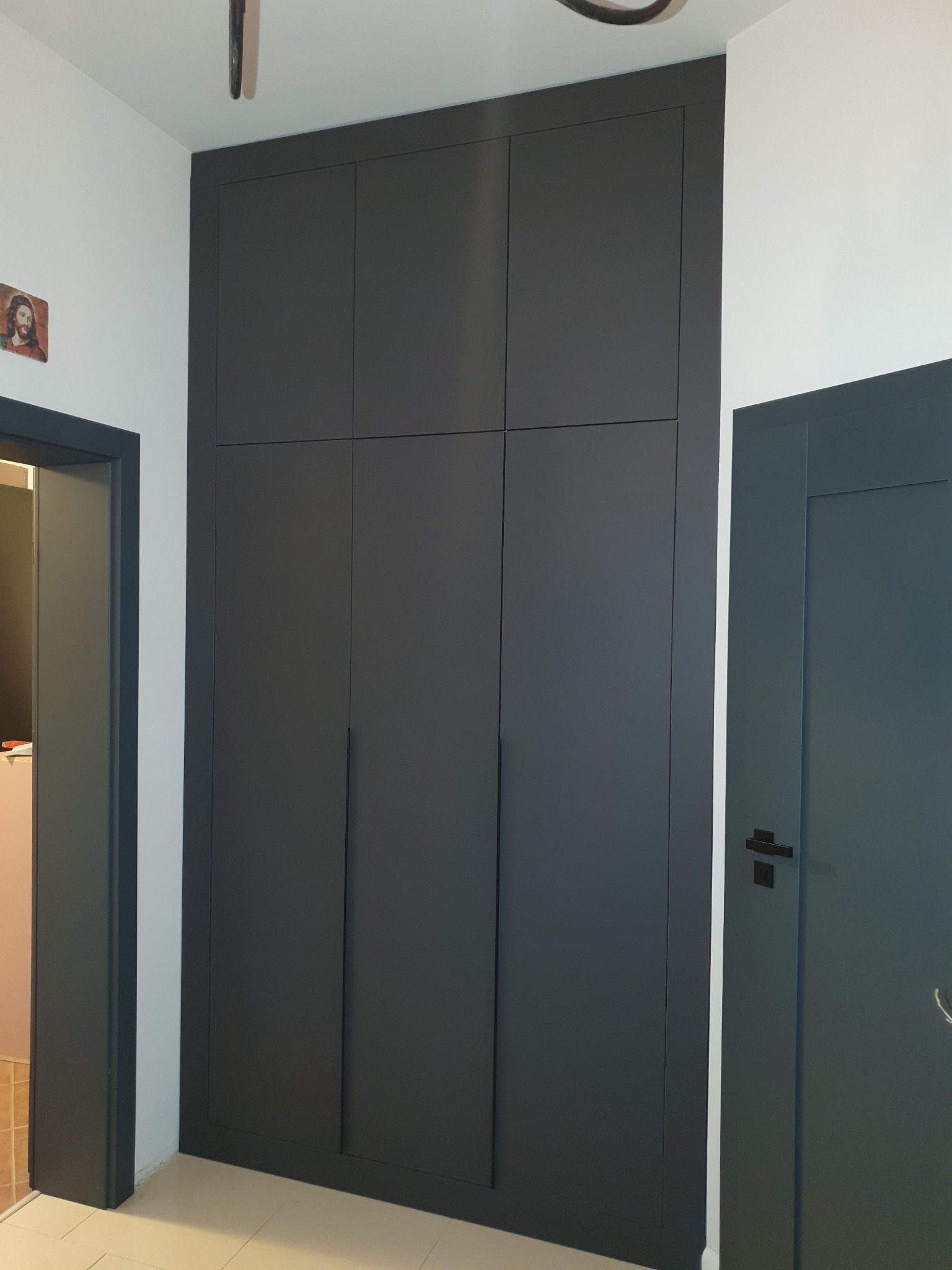 Wysoka, zabudowana szafa w kolorze grafitowym z sześcioma drzwiami, wkomponowana w przedpokój z płytkami podłogowymi i dwiema parami drzwi w ciemnych odcieniach szarości.