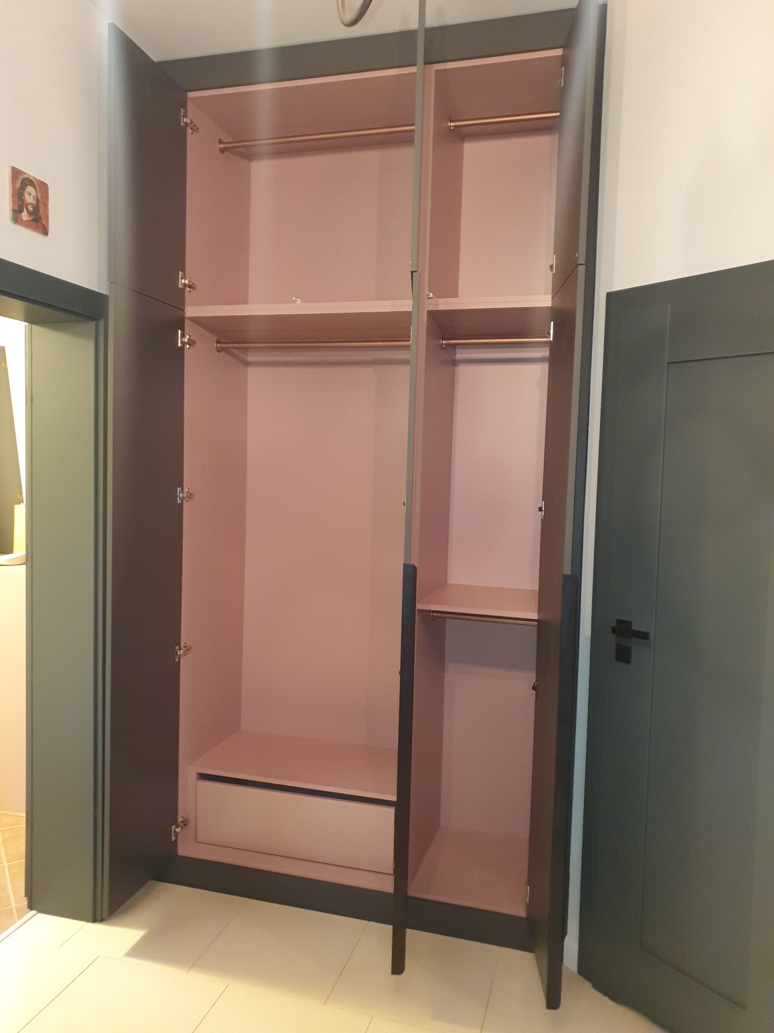 Otwarta szafa wnękowa z szarymi drzwiami i jasnoróżowym wnętrzem, widoczne półki, drążki na ubrania i szuflada, na ścianie obok drzwi wisi obrazek.