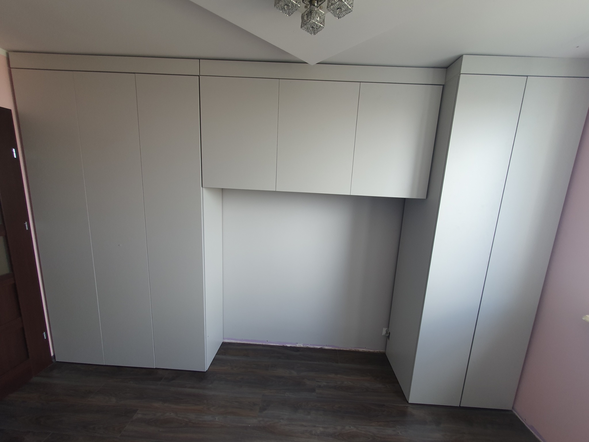 Jasnoszara zabudowa meblowa od podłogi do sufitu z miejscem na biurko, widoczne drzwi po lewej i drewnopodobna podłoga.