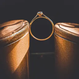 Złoty pierścionek zaręczynowy z brylantem ustawiony między dwoma złotymi szpilkami na ciemnym tle.