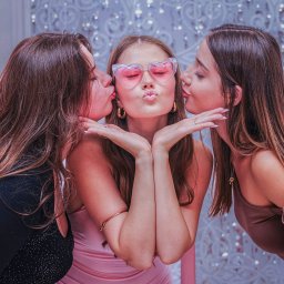 Piotr Pawlak_Fotografia Ślubna - Trzy kobiety w sukienkach, środkowa w okularach w kształcie serc, całowana w policzki przez dwie pozostałe na tle błyszczącej ściany.