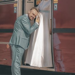 Piotr Pawlak_Fotografia Ślubna - Uśmiechnięty mężczyzna w błękitnym garniturze przykłada ucho do brzucha kobiety w jasnej sukni, stojącej w drzwiach czerwonego tramwaju. Nietypowa sesja ciążowa.