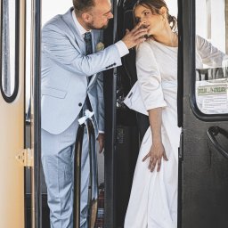 Piotr Pawlak_Fotografia Ślubna - Para młoda w tramwaju: pan młody w błękitnym garniturze dotyka twarzy panny młodej w białej sukni. Nietypowa sceneria ślubna z gaśnicą w tle.
