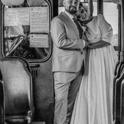 Piotr Pawlak_Fotografia Ślubna - Czarno-białe zdjęcie uśmiechniętej pary młodej w tramwaju Ikarus. Panna młoda w okularach w kształcie serc opiera głowę na ramieniu pana młodego.