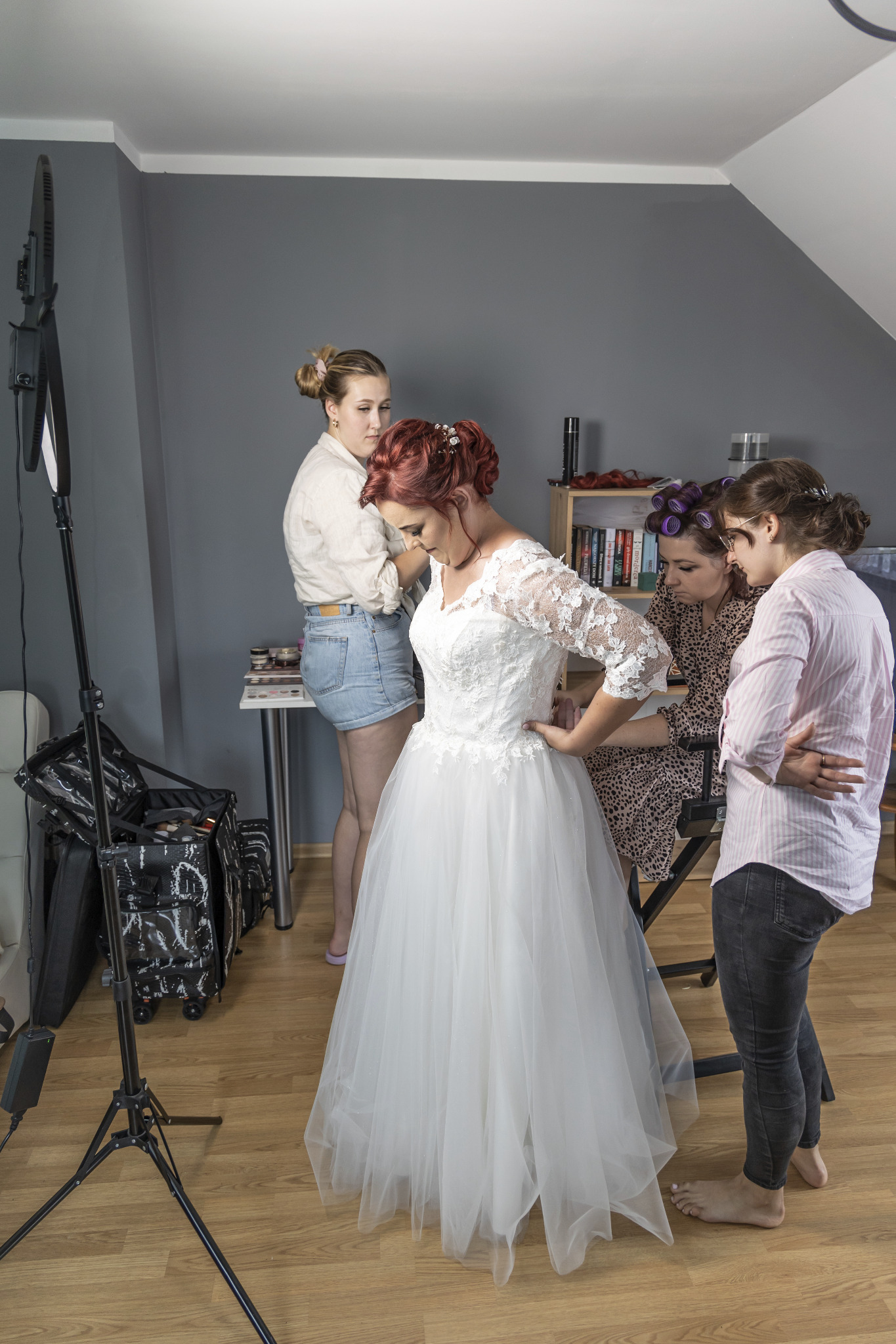 Panna młoda w białej sukni ślubnej przymierzana przez dwie osoby, trzecia osoba poprawia makijaż, w tle widoczny sprzęt oświetleniowy i kosmetyki.