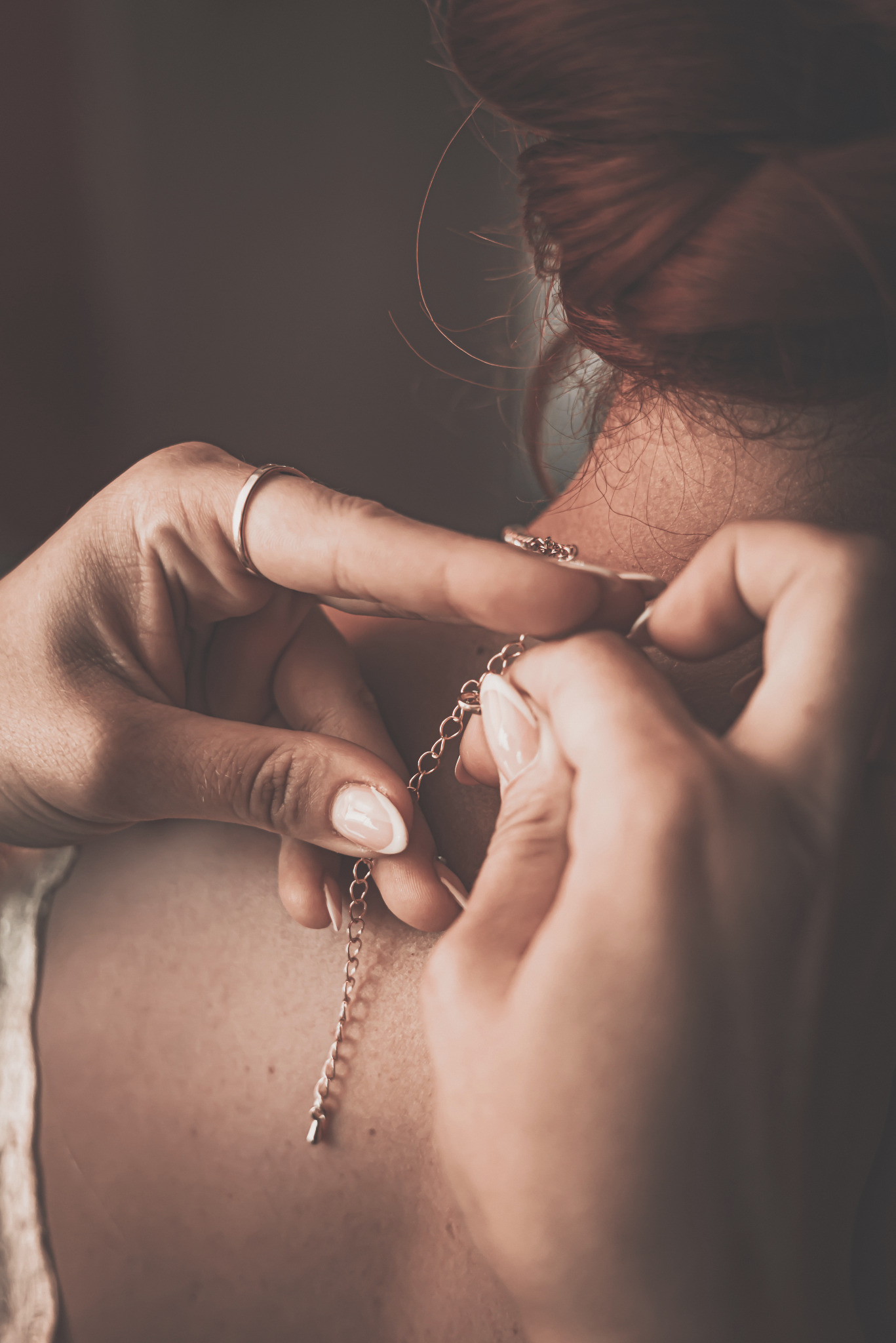 Dłonie kobiety z pierścionkiem pomagają zapiąć delikatny, złoty łańcuszek na szyi, widoczny kark i upięte rude włosy.