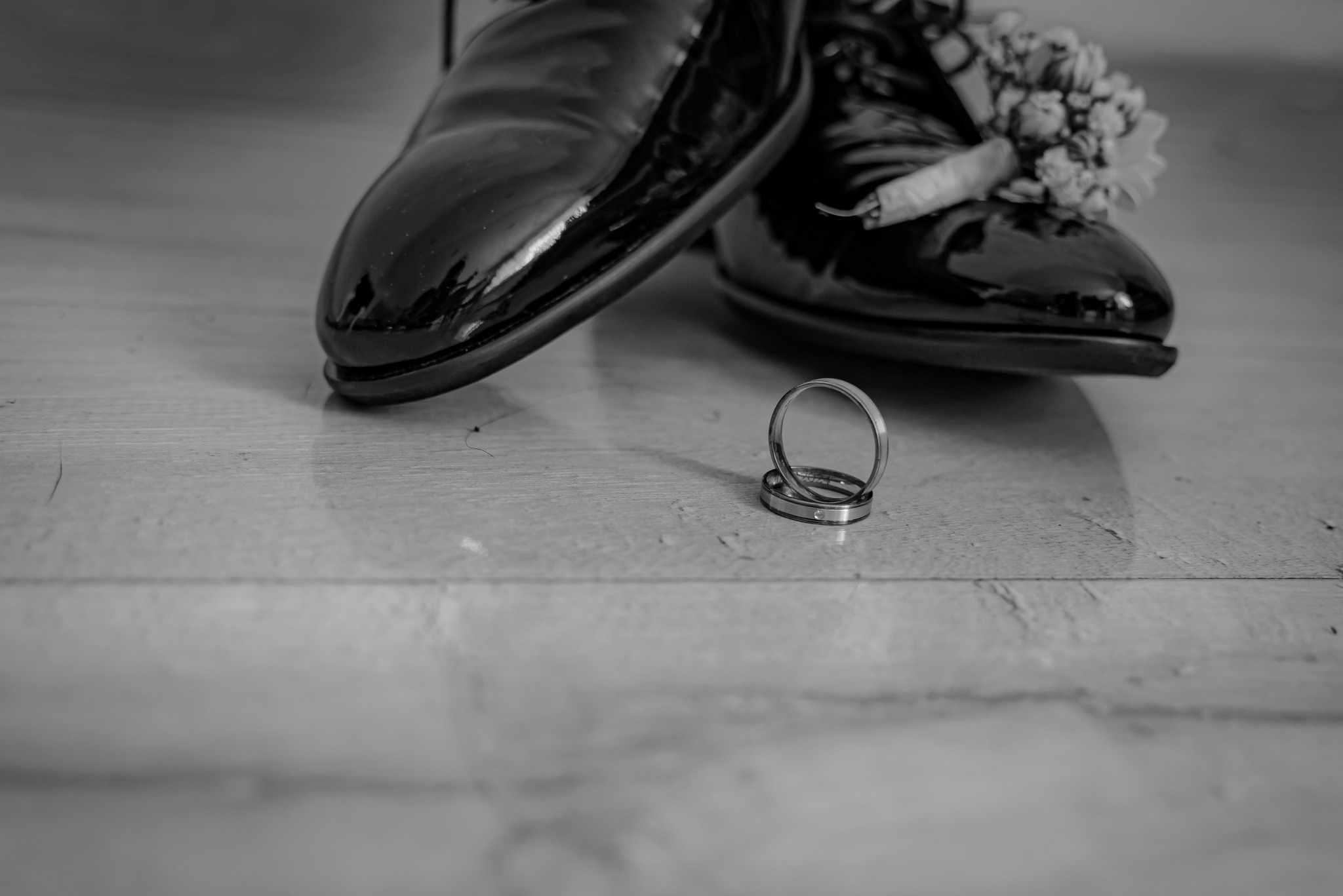 Czarne lakierki męskie z butonierką i obrączki ślubne na drewnianej podłodze w czerni i bieli.